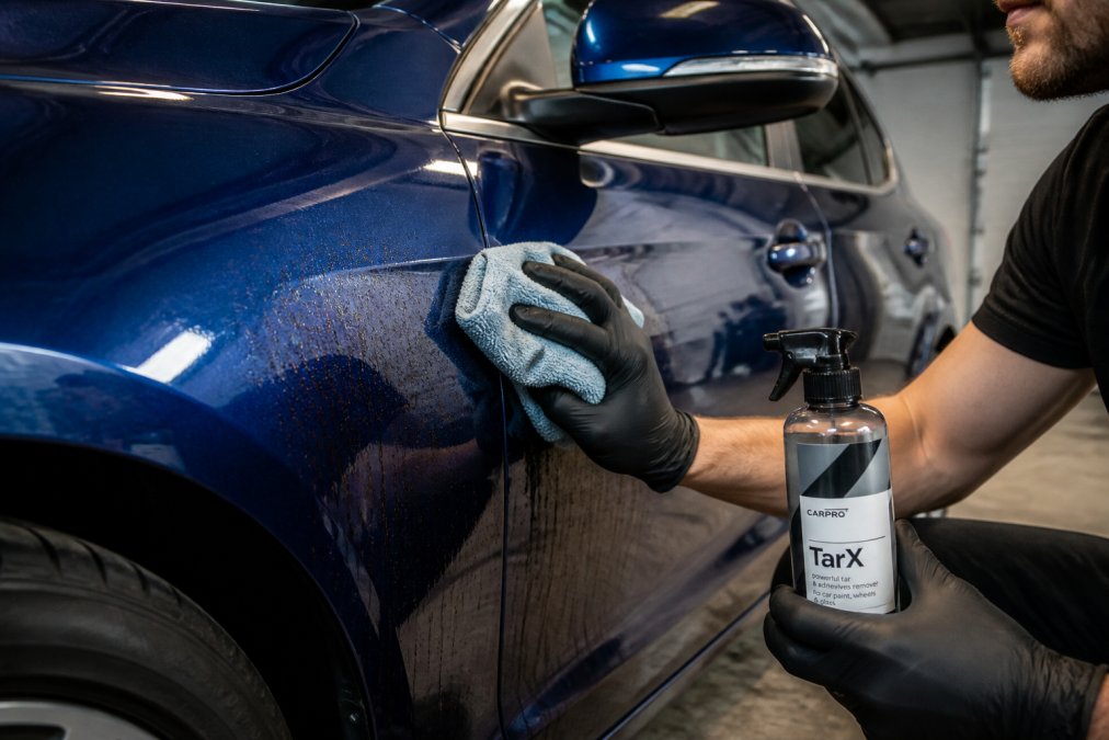 Professional demonstrating Carpro Tar.X application on dark blue vehicle using microfiber cloth with visible tar residue on automotive paint in natural garage lighting