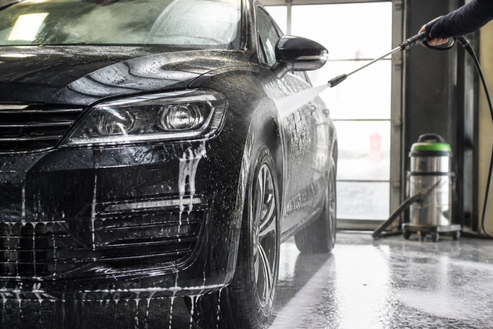 Vehicle in garage being washed with water and automotive shampoo post-tar removal, clean glossy paint visible under bright workshop lighting