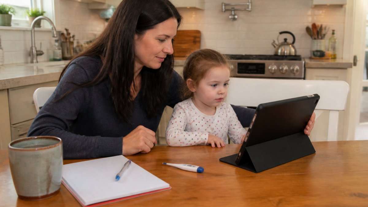A parent sitting at a kitchen table with a young child, holding a tablet propped up for a video call, with a thermometer and a notepad nearby