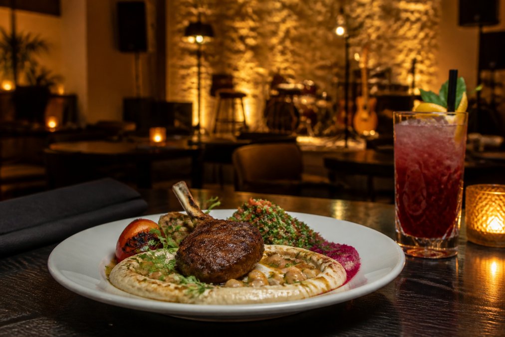 An elegantly plated Mediterranean dish on a dark wooden table with warm amber ambient lighting, a handcrafted cocktail glass nearby, and a softly lit stage visible in the background with subtle live music decor