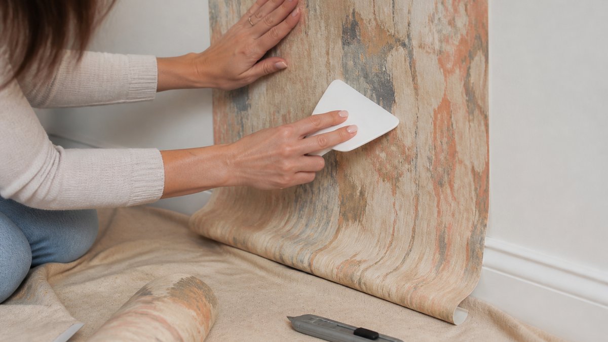 Close-up of a renter's hands smoothing a peel-and-stick wallpaper panel onto a clean interior wall, with a plastic smoother and utility knife on a nearby drop cloth
