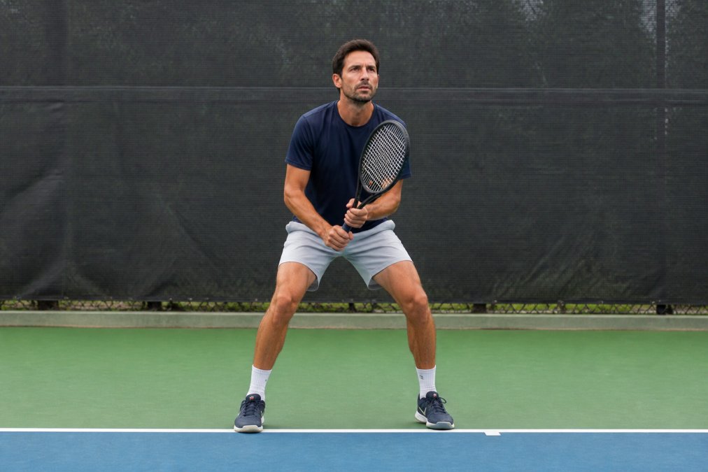 Adult tennis player demonstrating the split step position on a tennis court, feet shoulder-width apart, knees slightly bent, weight on the balls of the feet, ready to move in either direction with athletic posture