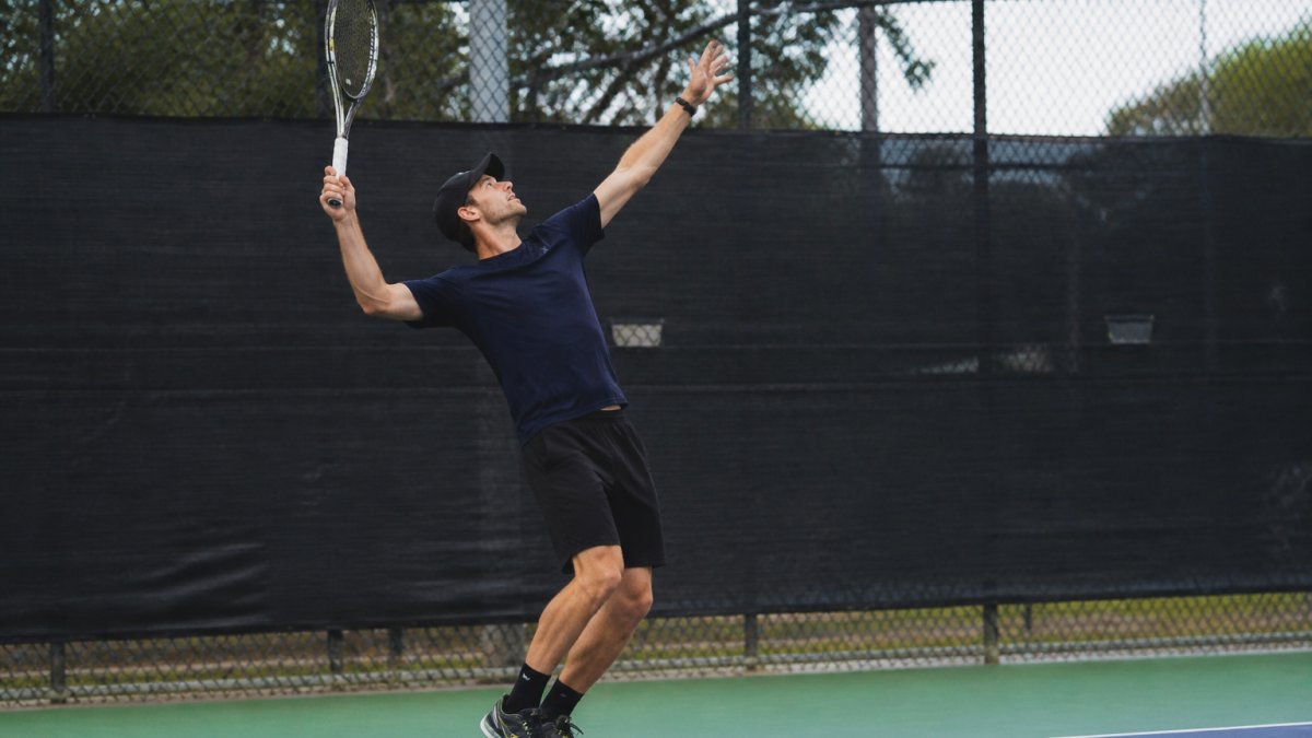 Recreational tennis player mid-serve captured from the side showing full body position during acceleration phase with racquet raised and legs engaged, natural outdoor court lighting
