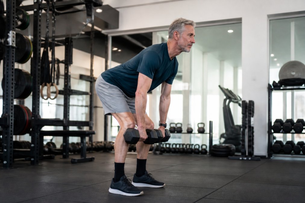 Mature male tennis player performing a Romanian deadlift with dumbbells in a well-lit gym, demonstrating controlled hip hinge movement and neutral spine positioning