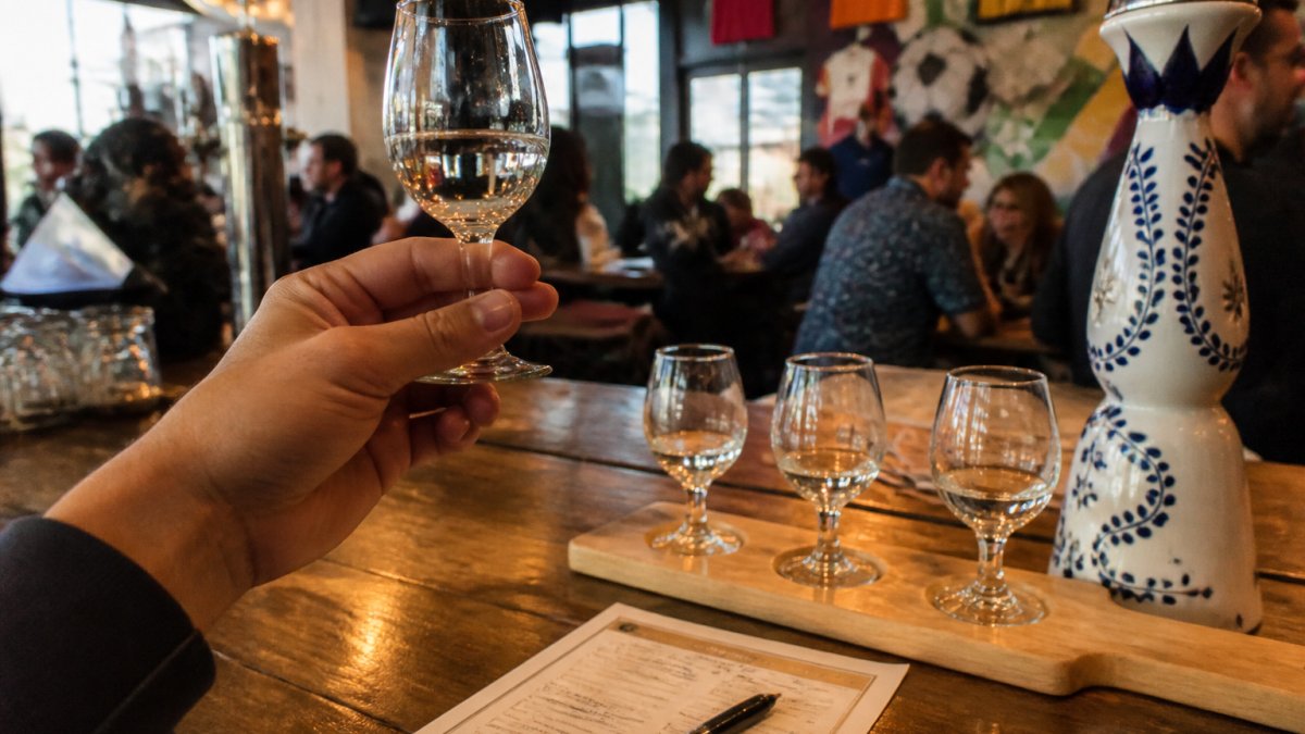Close-up of a person holding a small stemmed tasting glass of clear tequila up to the light, with a flight of three glasses and a tasting worksheet on a wooden bar top in a warm, lively cantina setting