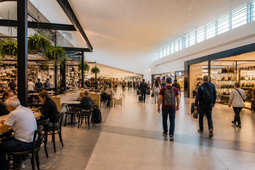 Modern retail and dining area inside Terminal 3 with cafes, shops, and passengers browsing or sitting with refreshments, natural lighting from large windows