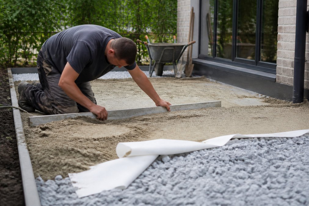 Vakman die drainage- en stabilisé-lagen aanlegt onder een terras, met zandmengeling, gereedschap en waterdoorlatingsmateriaal zichtbaar in de voorbereiding