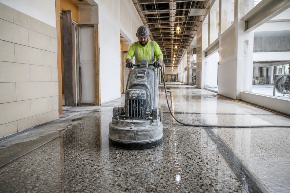 A commercial terrazzo installer operating a heavy grinding machine across a freshly poured terrazzo floor in a large institutional corridor, with aggregate chips and divider strips visible in the wet surface under bright construction lighting