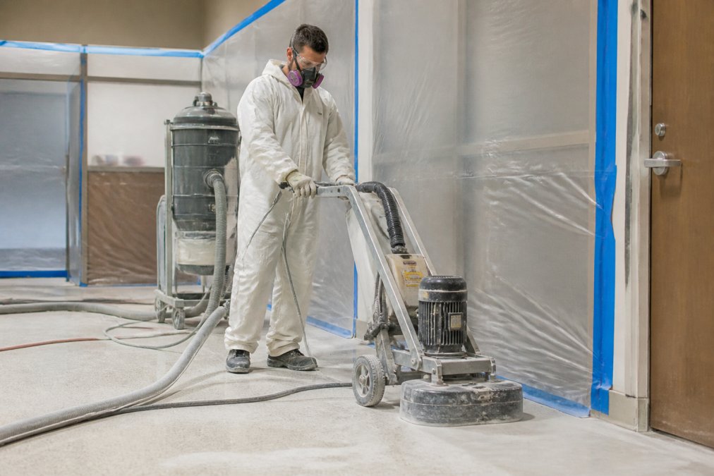 Worker in full protective equipment including respirator and coveralls performing substrate grinding with dust containment barriers and HEPA filtration system visible in healthcare facility preparation area