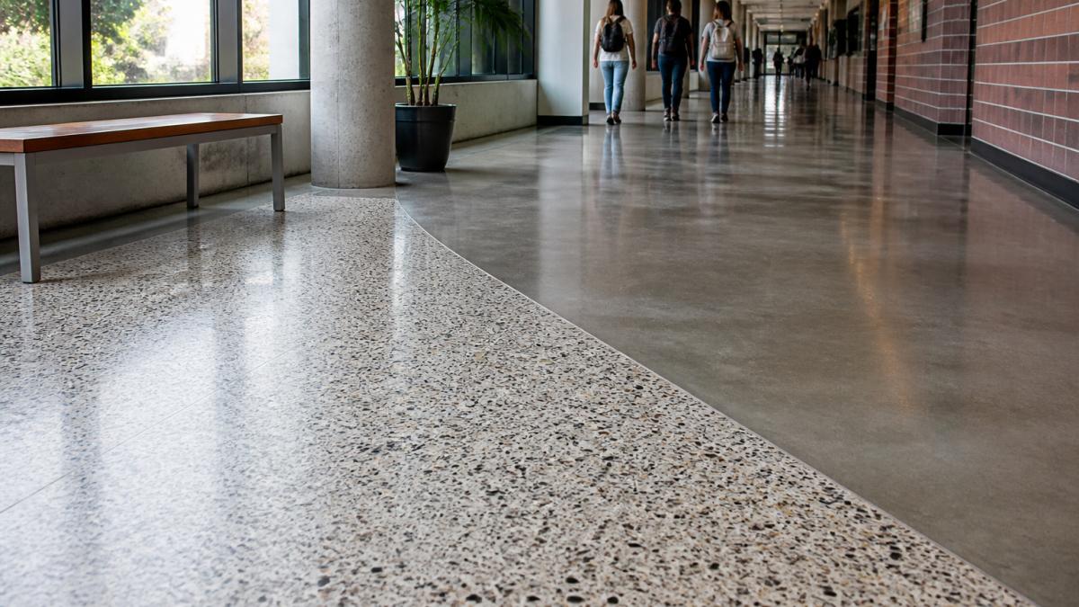 Wide-angle view of a modern university corridor with a terrazzo floor featuring integrated aggregate design in the foreground transitioning to a polished concrete section in the background, highlighting visual and textural differences between materials