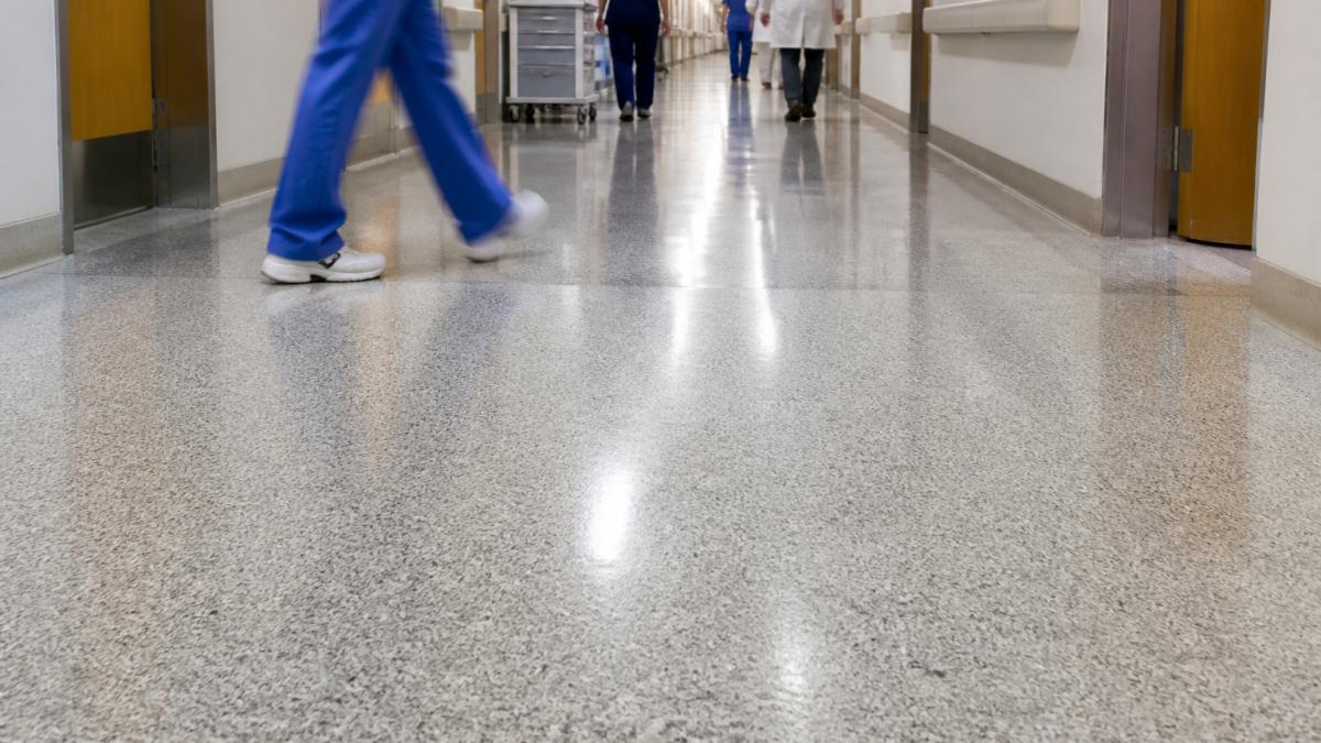 Busy hospital corridor with polished flooring surface showing foot traffic patterns, healthcare workers walking across the floor under bright clinical lighting