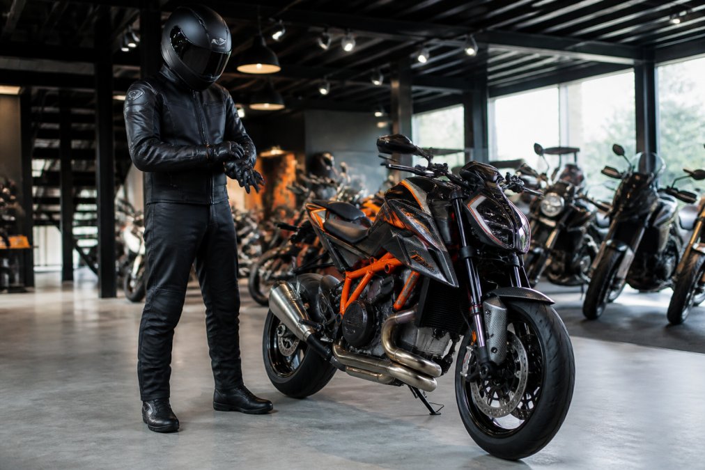 Motorrijder in volledige veiligheidsuitrusting met zwarte leren jas, integraalhelmmet, gevoerde motorhandschoenen en laarzen, staande naast een glanzende exclusieve motorfiets in een moderne dealerruimte met heldere verlichting