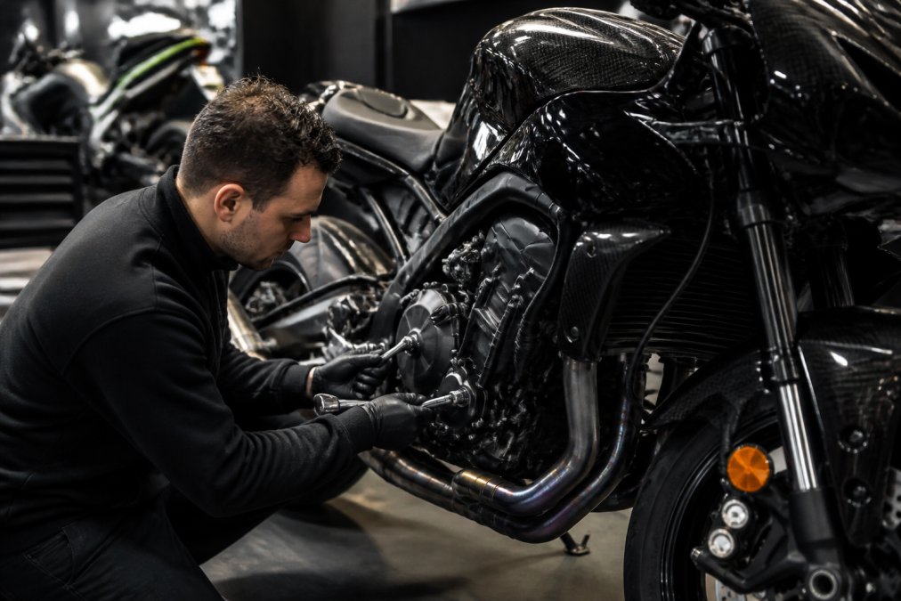 Technicus in werkkleding voert nauwgezette inspectie uit op de motorblok en prestatie-onderdelen van een glanzende exclusieve motorfiets, controlerende schroeven en leidingen met professionele gereedschappen in een heldere werkplaats