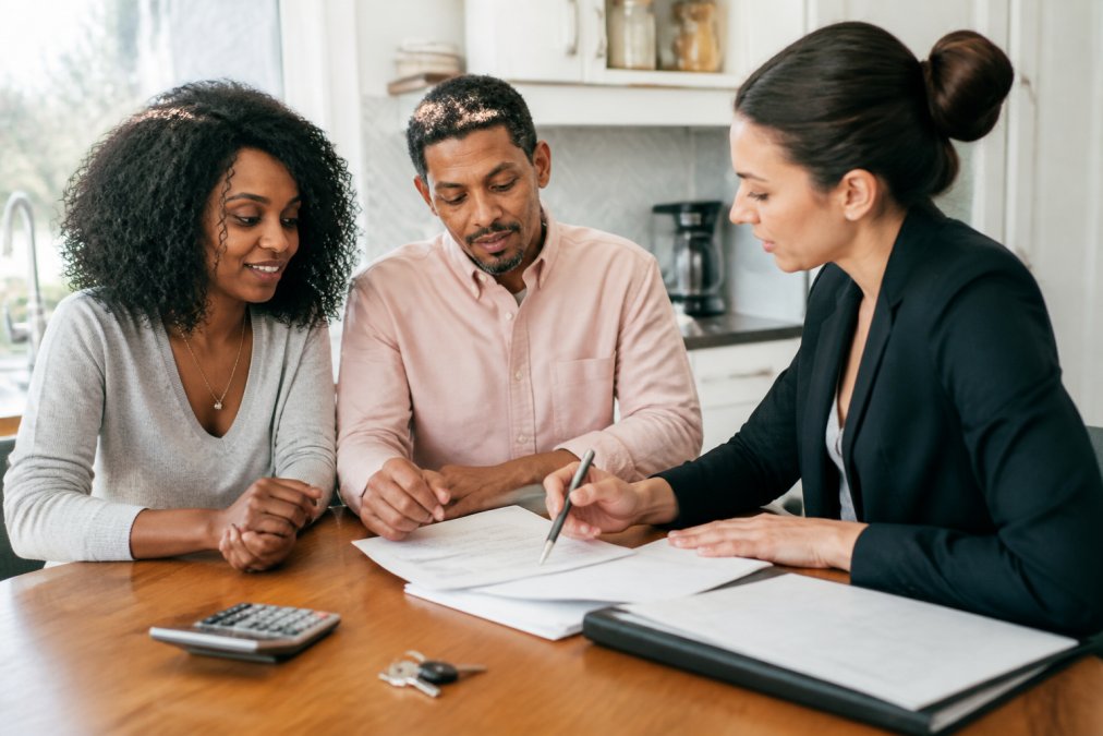 A couple reviewing financial documents with a real estate agent at a kitchen table, a house key and calculator beside a stack of papers, bright natural light filling a home office