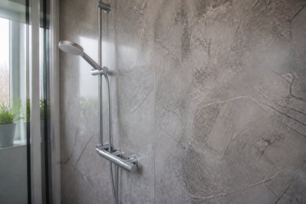 Close-up of a modern bathroom wall covered in textured stone-effect grey panels, with a chrome shower fixture and soft natural light from a window