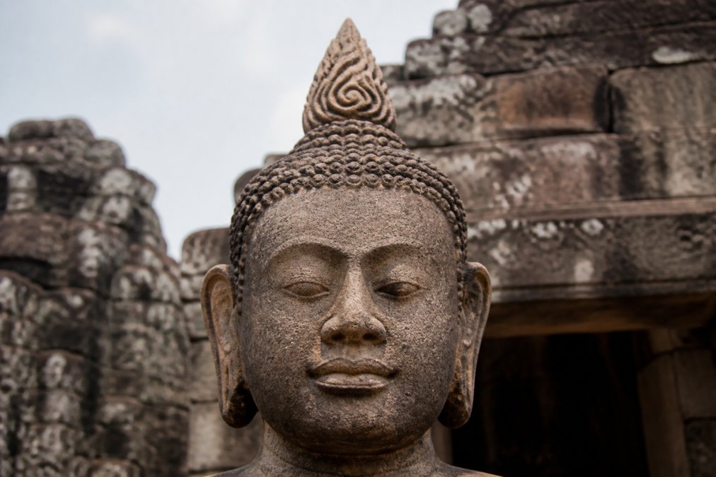 Close-up of an ancient Khmer sandstone Buddha statue showing the broad face, full cheeks, serene expression, and distinctive flame-like ushnisha finial carved into weathered stone