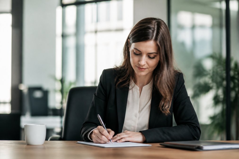 Professional woman writing a thank you note after job offer with pen and paper at a sunlit office desk.