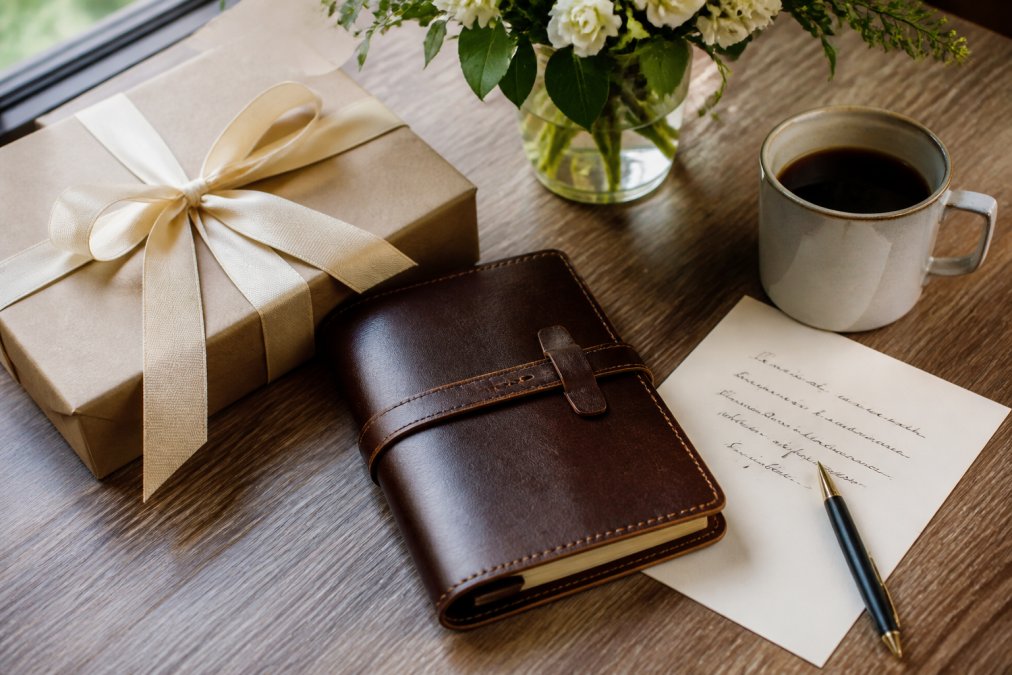 Carefully wrapped premium gift box with personalized leather journal and handwritten thank you note on polished desk with pen, coffee cup, and flowers in soft natural lighting