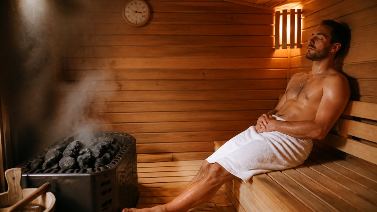Person relaxing on a wooden bench in a steamy sauna with a wall thermometer visible nearby