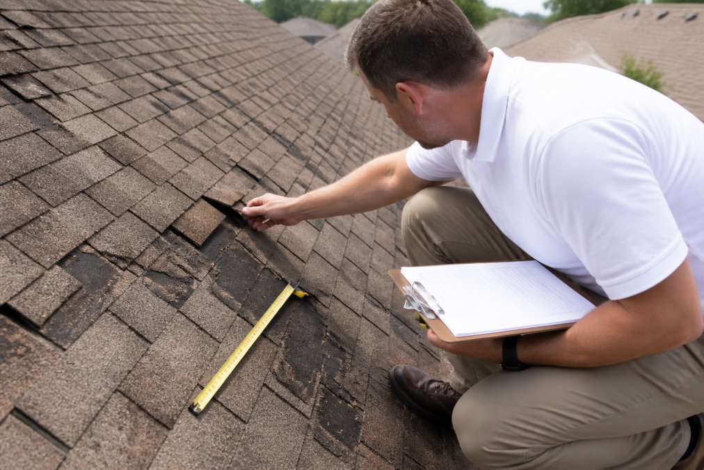Professional roofing inspector examining damaged shingles and roof sections with measuring tape and clipboard during daylight inspection