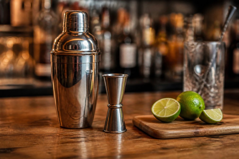 Close-up of a stainless steel cocktail shaker next to a jigger and fresh cut limes on a wooden bar counter, warm ambient lighting