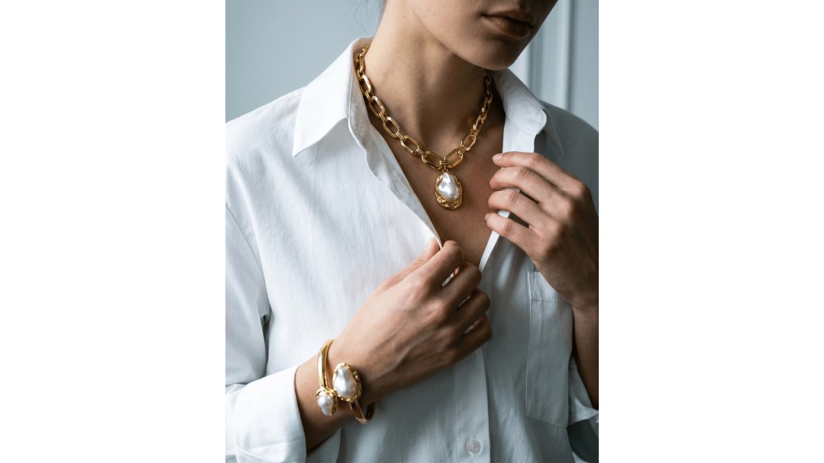A woman in a simple cream-colored top and jeans wearing a chunky gold chain necklace with a large baroque pearl pendant, standing in a bright boutique with warm natural light