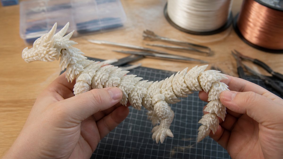Hands gently flexing the tail and leg joints of a finished 3D-printed articulated dragon on a workbench.