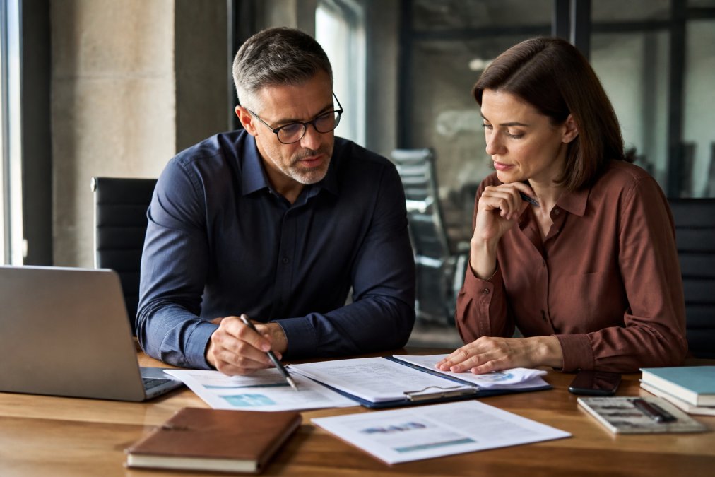 Business owner and consultant collaborating at a desk with marketing documents, laptop, and strategic plan visible, natural office lighting