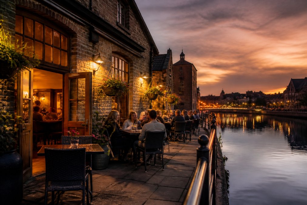 A warm, inviting exterior shot of an independent riverside wine bar with outdoor seating along the water in a historic English city at dusk, lights glowing from the windows