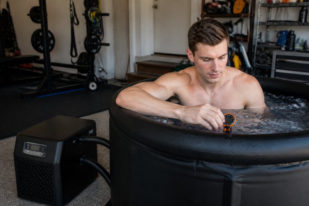 A person sitting in a portable ice bath tub in a home garage gym, checking a waterproof timer on the edge of the tub, with a water chiller unit nearby