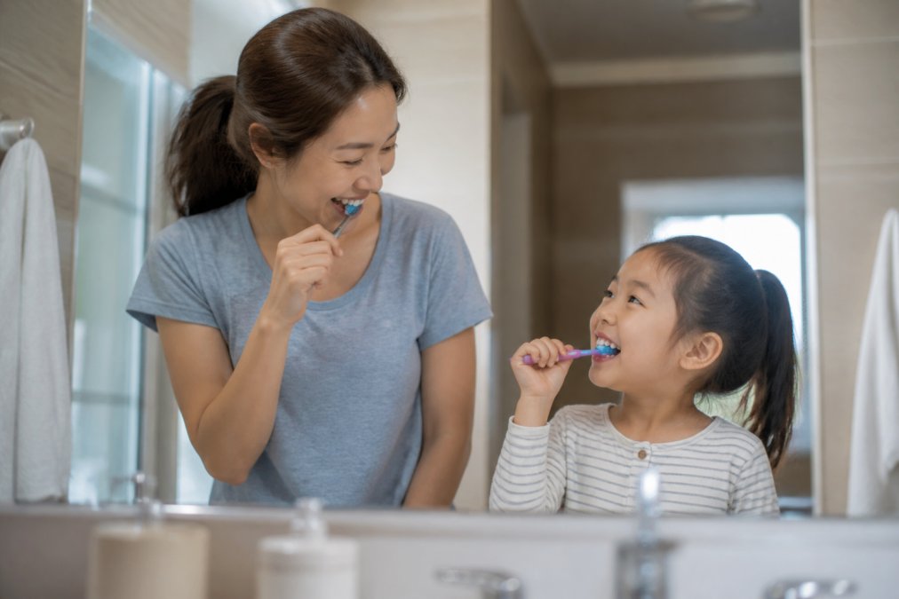 A parent and young child standing side by side at a bathroom sink, both brushing their teeth in a bright, tidy home bathroom, smiling at each other in the mirror