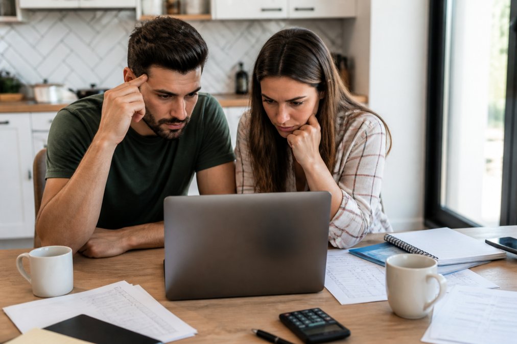 A couple sitting at a bright kitchen table, both looking at a single laptop screen showing a wedding planning layout, with printed checklists and coffee mugs nearby