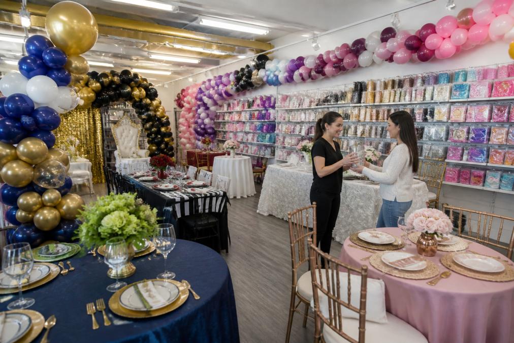 Inside a vibrant party store showroom with colorful balloon arches and arrangements displayed on walls, themed table settings with linens and centerpieces, shelves stocked with party supplies organized by category, and a friendly staff member helping a customer select decorations