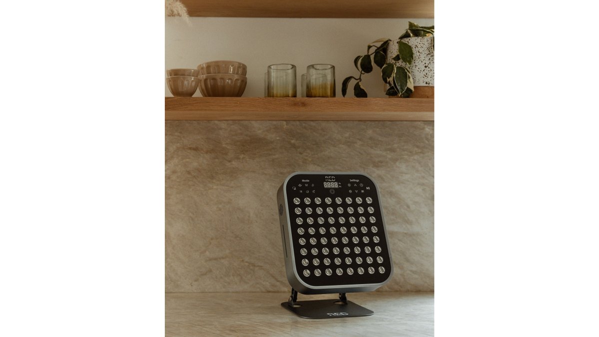 A neatly organized corner of a modern apartment with a folded foam roller, a small red light therapy panel on a shelf, and a rolled-up massage mat leaning against the wall