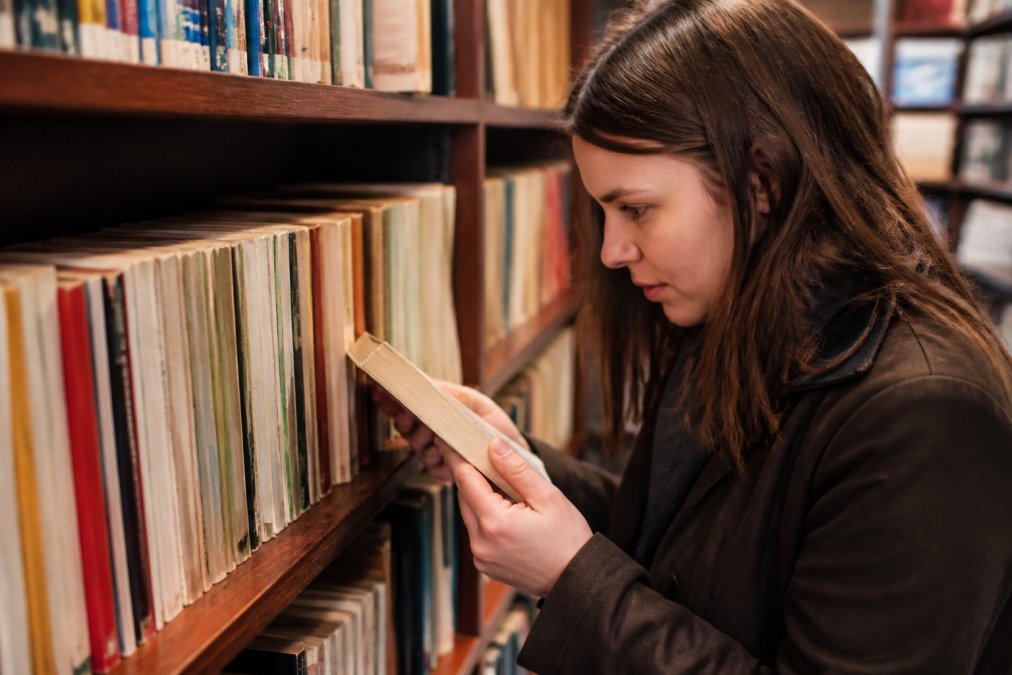 Person carefully examining used paperback books on a shelf, checking spine condition and page quality before making a selection