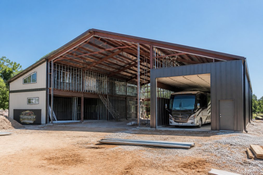 A barndominium under construction with a large attached shop space, steel frame exposed, open floor plan visible, and an RV parked in the wide garage bay under clear daylight