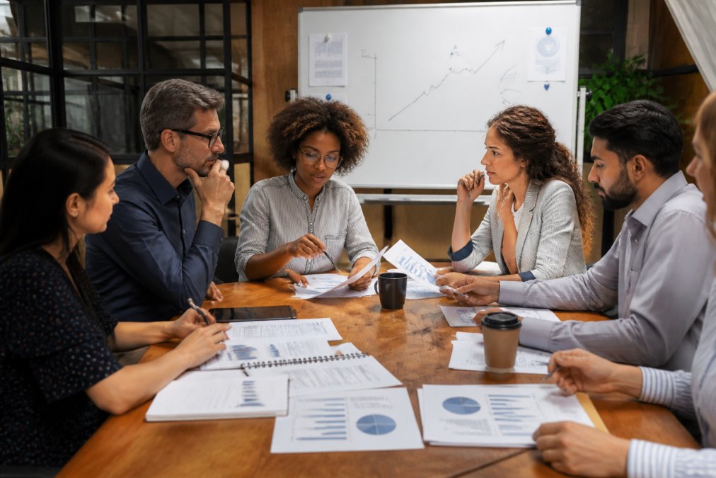 A diverse team of marketing professionals gathered around a conference table, comparing agency proposals on laptops and printed reports, with a whiteboard showing a growth chart in the background