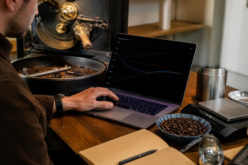 Coffee roaster monitoring a laptop screen displaying thermal roast profile curves and real-time bean temperature data, with a cup of freshly roasted coffee beans and roasting equipment visible nearby