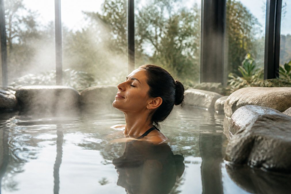 Guest relaxing in a warm thermal pool with steam rising, natural daylight filtering through windows, facial expression showing deep calm and restoration
