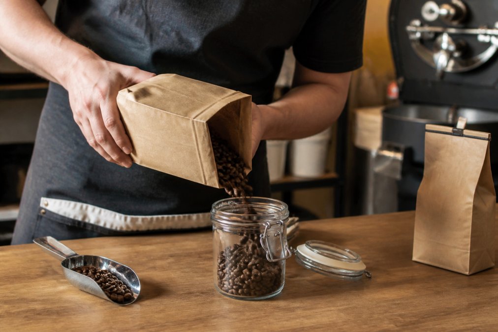 Person pouring fresh roasted coffee beans from a kraft paper bag with roast date label into a glass storage jar on a wooden counter