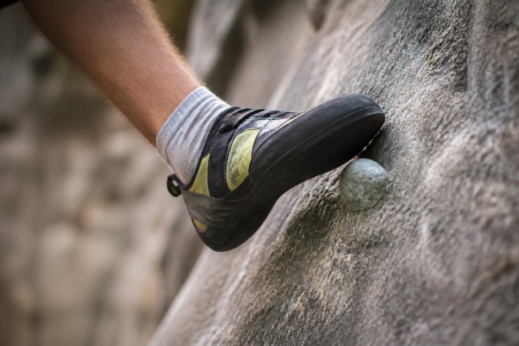 Close-up of a climber's foot in a tight climbing shoe gripping a small textured rock hold, showing the toe box pressed against the wall with clear contact points