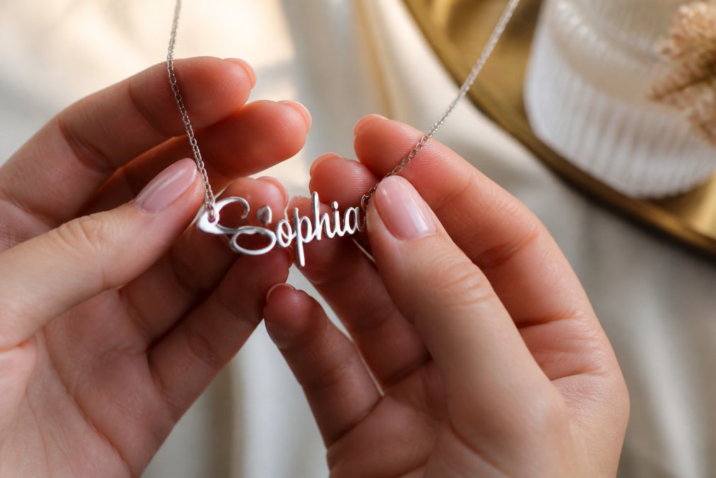 Close-up of hands holding a personalized name necklace with cursive engraving, polished stainless steel catching soft window light, delicate cable chain draped between fingers
