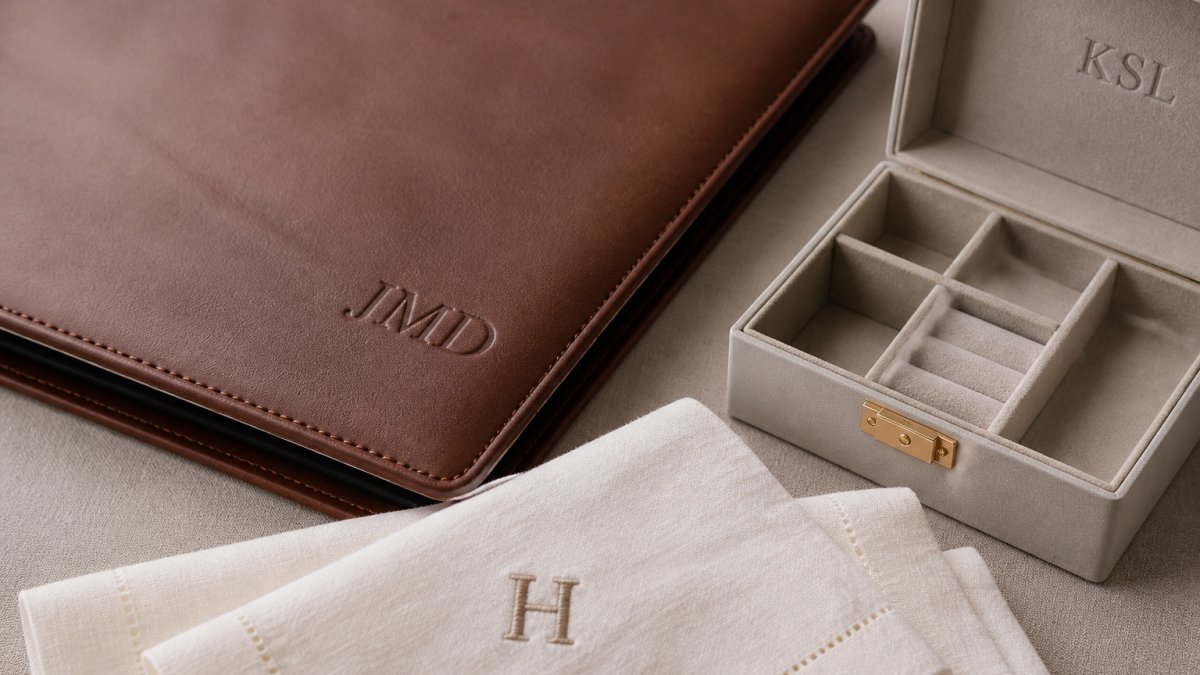 Close-up of personalized monogrammed items including engraved leather portfolio, custom jewelry box, and monogrammed linen napkins arranged on a neutral surface, showing fine detail of engraving craftsmanship and material texture