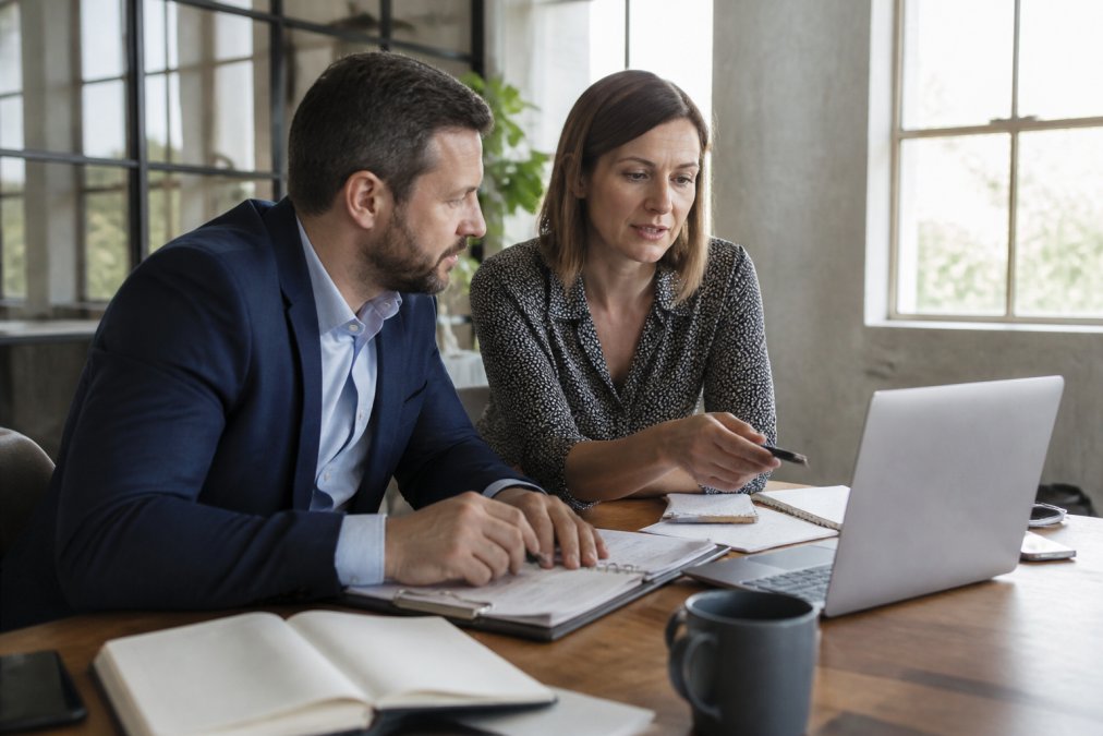 Professional consultant and business owner reviewing sales process workflow on laptop at modern office desk with notebooks and strategy documents visible