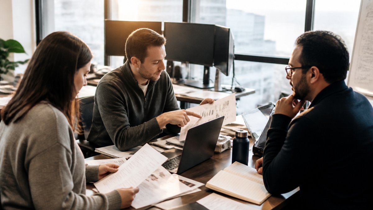 Security team members in modern office reviewing vendor risk assessments and monitoring dashboards on multiple computer screens, collaborating around a desk with laptops and compliance documents