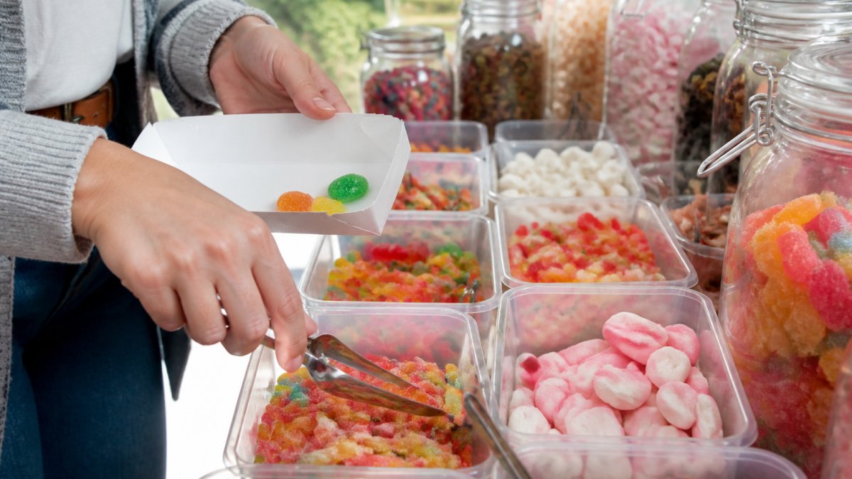 Close-up of hands selecting colourful sweets from a pick and mix selection, with various jars and containers of gummies, jellies, and chocolates visible in natural daylight