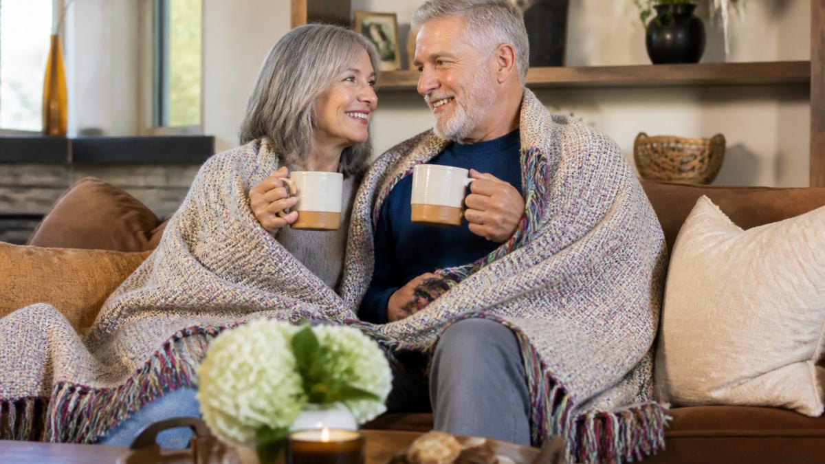An older couple sitting together on a couch, wrapped in a cozy woven cotton blanket with colorful fringe, smiling and holding matching coffee mugs in a warm living room