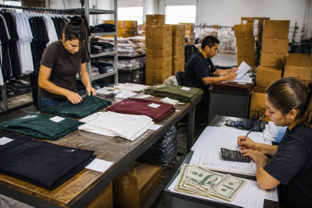 Busy operations center with staff reviewing custom apparel orders, quality control samples laid out on inspection tables, with printed garments hanging on racks and shipping boxes stacked in background
