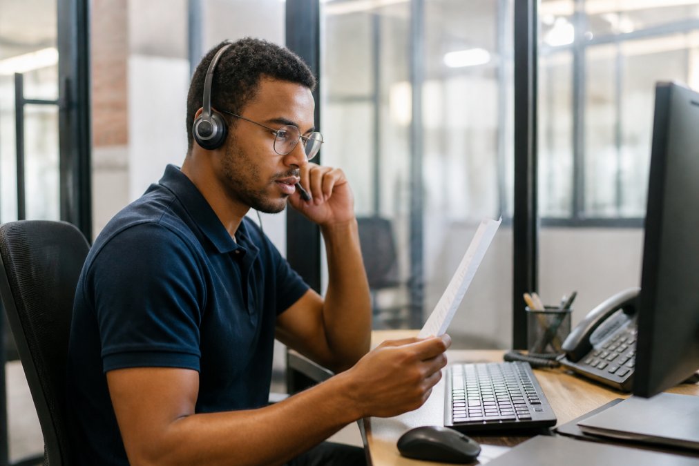 Help desk technician wearing a headset at a workstation, assisting a customer via phone while reviewing technical documentation on a second monitor, with ticket management software visible in the background