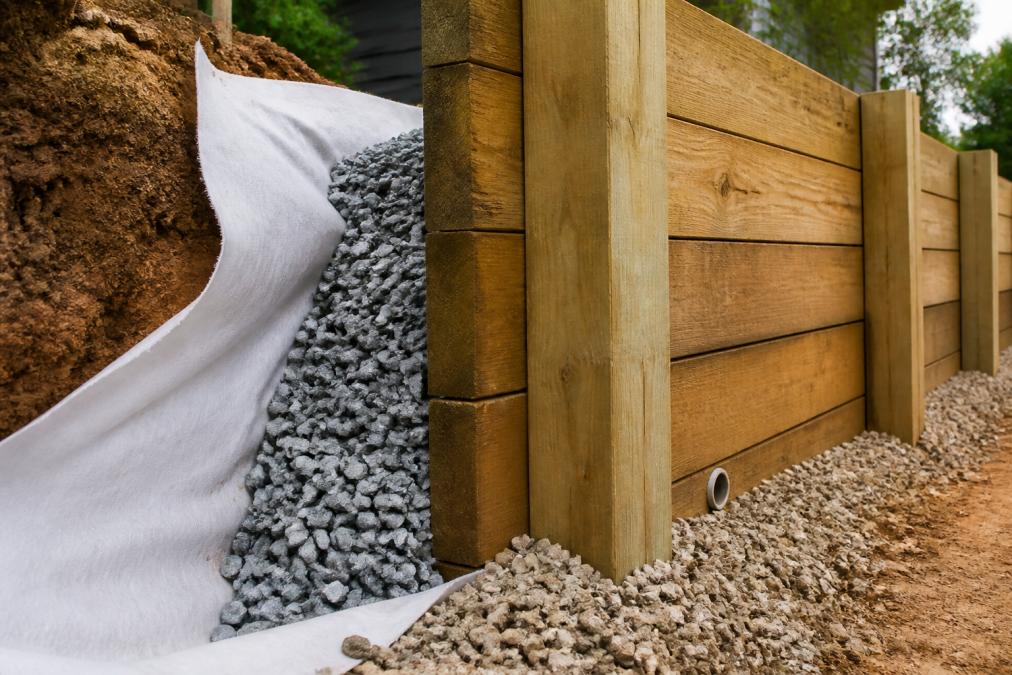 Cross-section showing drainage aggregate and geotextile fabric positioned behind a timber retaining wall with visible weep holes at the base allowing water drainage